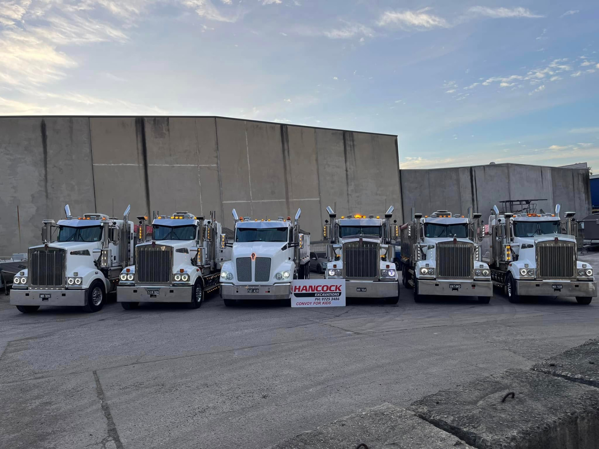Hancock Excavations trucks lined up at the yard behind a Convoy for Kids sign in 2023