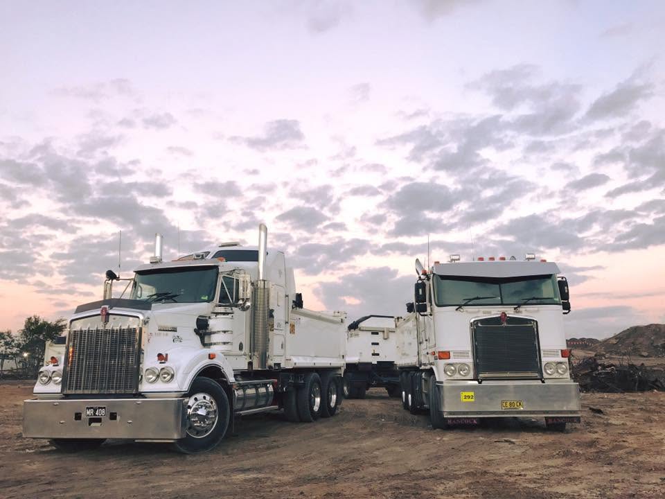 Two Hancock Excavations tippers, including a truck-and-dog combination, parked on a dirt work site at dusk